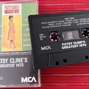 May include: A vintage cassette tape and case for Patsy Cline's Greatest Hits. The case features a photo of Cline in a yellow dress. The black cassette tape has the album title and song titles printed on it. The background is red.