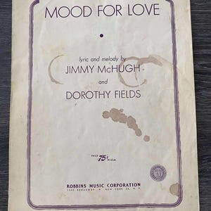 May include: A vintage sheet music cover for "I'm in the Mood for Love." The cover displays the title in large, bold letters, with the lyricist and composer's names below. The cover is cream-coloured with a purple border and some stains.