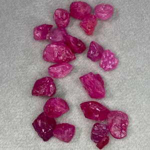 May include: A collection of raw, uncut ruby gemstones. The stones vary in size and shape, displaying a vibrant pink to deep red hue. The rough texture and natural forms of the rubies are visible against a neutral background.