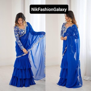 Puede incluir: Un sari azul vibrante con intrincados bordados plateados en la blusa de manga larga y el borde. El sari presenta una falda con volantes en capas y una caída transparente y fluida. El texto "NikFashionGalaxy" se muestra en un rectángulo negro.