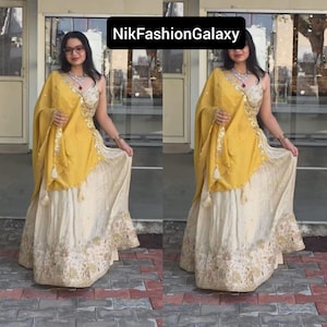 May include: A two-piece cream and yellow ethnic outfit. The top is a cream-colored, sleeveless, embellished top. The skirt is cream-colored with gold embroidery. A yellow shawl is draped over the shoulder. The text "NikFashionGalaxy" is at the top.