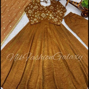 Puede incluir: Un traje Anarkali marrón dorado con bordados florales en el corpiño y las mangas. El conjunto incluye una dupatta y un pantalón a juego. El texto "Nik Fashion Galaxy" es visible en el vestido.