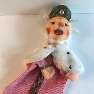 May include: A pink corduroy puppet with a white fur collar and a gold trim. The puppet has a white beard and a painted face with a crown on its head. The crown is teal blue with gold trim and a pearl.