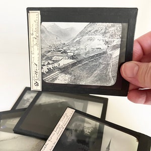 May include: A set of vintage glass lantern slides featuring black and white photographs of a Swiss town and a train travelling through a mountain pass. The slides are labelled with the text "Zurich from Quai Bridge" and "Scerne and Railway Train".