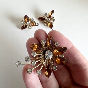 May include: A vintage jewellery set comprising a large brooch and a pair of earrings. The brooch is a gold-toned flower with brown and clear rhinestones. The earrings are also gold-toned and feature brown and clear rhinestones.
