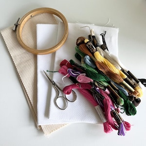 May include: A craft kit with embroidery supplies. The kit includes a wooden embroidery hoop, white and beige cross-stitch fabric, a pair of small scissors, and a variety of colourful embroidery floss. The floss colours include shades of pink, green, yellow, and brown.