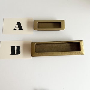 May include: Two antique gold cabinet door pulls. The top pull is smaller than the bottom pull. Both pulls have a rectangular shape with a recessed centre.