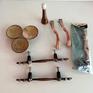 May include: A collection of hardware including three round brass-toned knobs, two twisted copper-toned drawer pulls, two long copper-toned handles with black accents, a conical door stop with a white tip, and one packaged handle labelled 715 AC.
