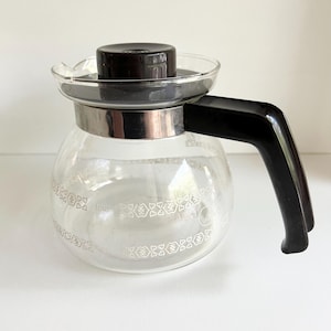 May include: A clear glass coffee carafe with a black lid and handle. The carafe has a silver band around the middle and a decorative white floral pattern. The carafe is designed for brewing coffee.
