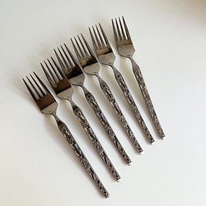 May include: Six stainless steel forks with a textured handle design. The forks have four tines and a dark gray finish, arranged diagonally on a white surface. Ideal for a formal dining setting.