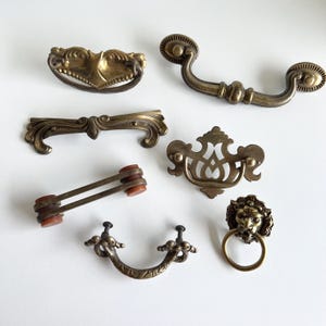 Könnte beinhalten: Eine Auswahl an antiken Messing-Schubladengriffen und -griffen in verschiedenen Stilen. Die Sammlung umfasst verzierte Designs, wie einen Löwenkopf-Ringgriff, gebogene Griffe und dekorative Stangengriffe. Die Hardware ist in Gold- und Brauntönen gehalten.