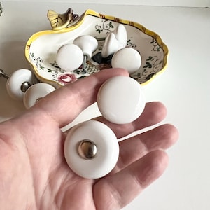 Peut inclure: Un ensemble de boutons de cabinet en céramique blanche avec des accents argentés. Les boutons sont ronds et ont une finition lisse.