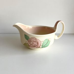以下が含まれることがあります： バラのデザインが施されたクリーム色のグレービーボート。ボートには注ぎ口、ハンドル、ピンク色の内装があります。ピンクのバラと緑の葉が側面に描かれています。グレービーボートはおそらくセラミックまたは磁器製です。
