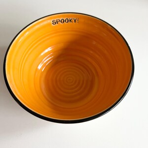 May include: Orange ceramic bowl with a black rim and a swirling pattern. The bowl has the word "SPOOKY!" written on the inside rim.