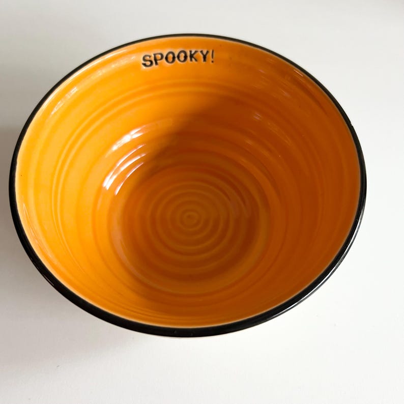 May include: Orange ceramic bowl with a black rim and a swirling pattern. The bowl has the word "SPOOKY!" written on the inside rim.