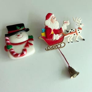 May include: Two Christmas brooches: a white snowman wearing a black top hat with a red and white candy cane, and a red sleigh carrying Santa Claus holding a sign that says NOEL, pulled by a white reindeer with black spots and a hanging metal bell.