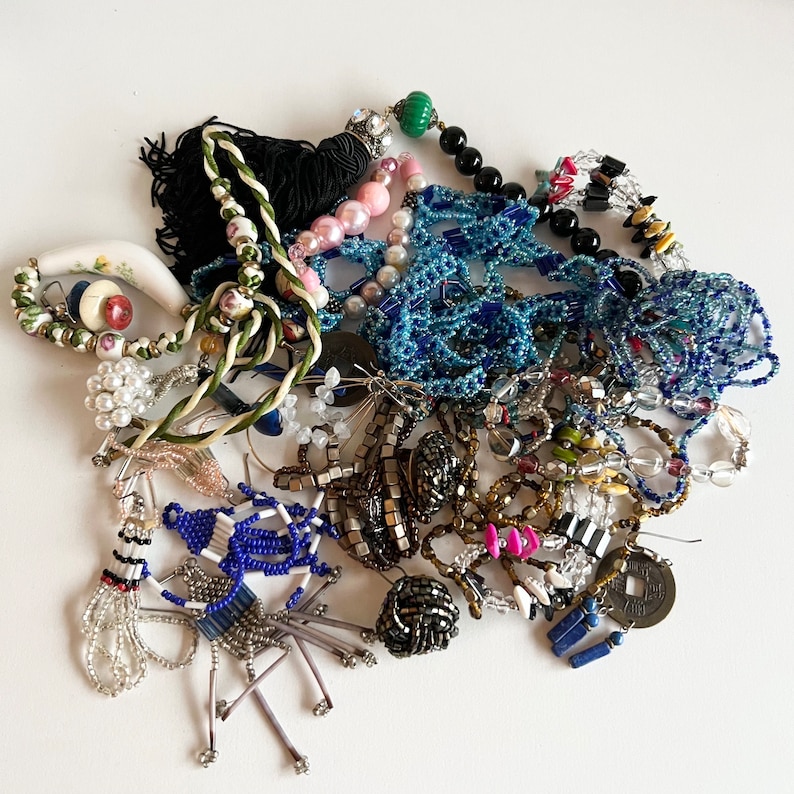 Puede incluir: Una colecci&oacute;n de joyas de cuentas de colores, incluyendo collares, pendientes y pulseras. Las joyas presentan una variedad de materiales, incluyendo cuentas de vidrio, cuentas de metal y cuentas de madera. Las joyas est&aacute;n dispuestas en una pila sobre una superficie blanca.