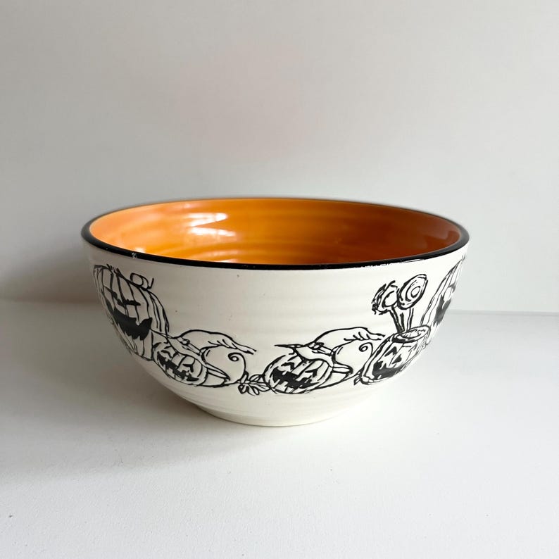 May include: A white ceramic bowl with a black and white Halloween design of pumpkins and bats. The inside of the bowl is orange.