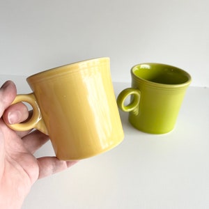 May include: Two ceramic mugs, one yellow and one green. The yellow mug has a handle and is being held in a hand. The green mug is sitting on a white surface.