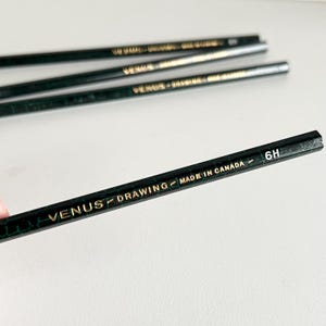 Peut inclure: Gros plan sur plusieurs crayons Venus Drawing verts avec des lettres dorées. Le texte sur les crayons indique "VENUS DRAWING MADE IN CANADA 6H". Les crayons sont disposés sur une surface blanche.