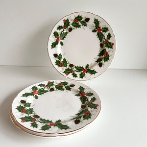 May include: Three white ceramic plates with gold rims. Each plate has a wreath design of green holly leaves, red berries, and brown pine cones. The plates are arranged on a white surface.