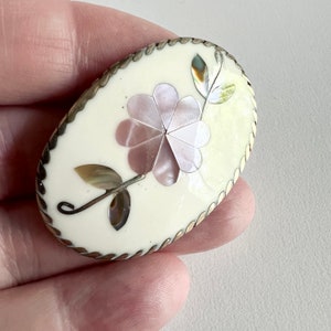 May include: Oval shaped brooch with a white background and a silver border. The brooch features a pink flower made of mother of pearl with a silver stem and leaves.