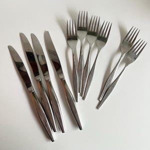 May include: A selection of stainless steel cutlery, comprising knives and forks, displayed on a white surface. The knives feature a streamlined, elongated form, and the forks have four prongs. This cutlery is suitable for dining.