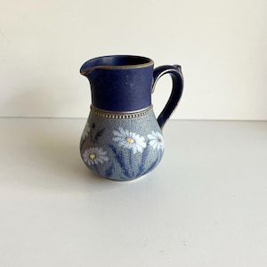 May include: A ceramic jug with a dark blue top and handle, and a grey body decorated with white daisies and gold centres. The jug has a spout and a gold band around the middle.