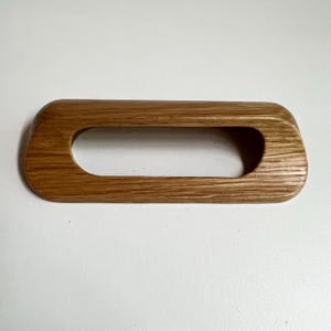 May include: A rectangular wooden drawer pull with a smooth, light brown finish and a visible natural wood grain pattern. The handle features a large, elongated oval cutout in the center, creating a hollow, open-frame design with rounded edges.