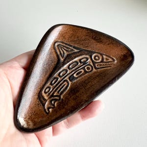 May include: A brown ceramic dish with a triangular shape and a carved design of a stylised salmon. The dish has a rustic, hand-crafted look.