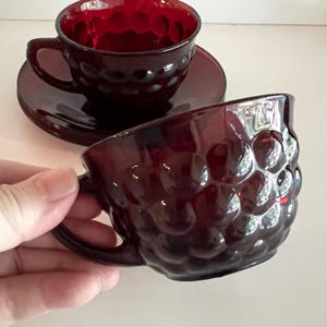 Peut inclure: Deux tasses en verre rouge profond avec un motif à pois en relief. Une tasse est placée au premier plan, tandis que l'autre repose sur une soucoupe en verre rouge assortie à l'arrière-plan. Le verre est translucide et présente un motif répétitif.
