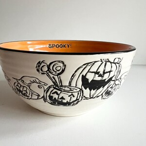 May include: A white ceramic bowl with a black rim and orange interior. The bowl has a black and white Halloween design with pumpkins, candy corn, and the word "SPOOKY!" on the rim.