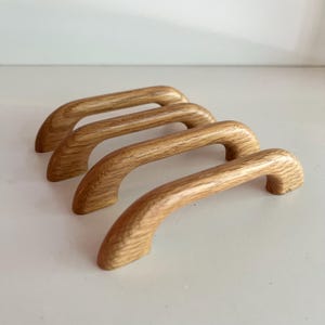 May include: Four arched cabinet handles crafted from light-toned wood with a visible natural grain pattern. Each handle features a smooth, curved design with two mounting points at the ends, showcasing a consistent light brown colour throughout.