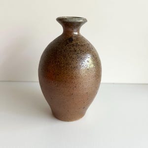 May include: A handmade, brown ceramic vase with a speckled, textured surface. The vase has a rounded body and a narrow neck with a slightly flared rim. The colour is a mix of brown and tan, with darker spots.