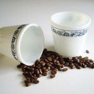 May include: Two white coffee cups with a blue floral pattern, one on its side, with scattered roasted coffee beans. The cups are cylindrical and feature a delicate floral band, ideal for coffee or espresso.