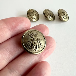 May include: A set of antique brass buttons. Each button features a crown above the letters "M" and "A". The buttons are round with a raised design. The buttons are a warm, antique brass colour.