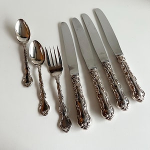 以下が含まれることがあります： スプーン、フォーク、ナイフを含むアンティークの銀食器セット。銀食器は、装飾的で詳細なハンドルと、光沢のある反射性のブレードとボウルを備えています。白い表面に配置されており、ヴィンテージカトラリーのエレガントなデザインを示しています。