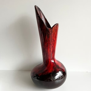 May include: A red and black ceramic vase with a unique, elongated, curved design. The vase has a glossy finish and a mottled glaze.