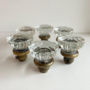 May include: A set of clear, ribbed glass door knobs with brass bases. The knobs have a flower-like design and are arranged on a white surface. The transparent glass allows light to pass through, creating a sparkling effect.