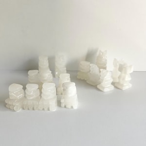 May include: A collection of white, carved chess pieces. The pieces are arranged in groups, with pawns, rooks, knights, and other pieces visible. The pieces have a smooth, polished appearance and are set against a plain white background.