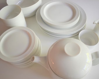 May include: A set of white porcelain dishes, including bowls, plates, a teapot, and a creamer. The dishes are stacked and arranged in a circular pattern on a white surface.