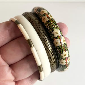May include: Four bangles in a variety of styles. One is ivory with gold-tone accents, another is a perforated metal, and the third is black with white floral designs. The bangles are stacked together.