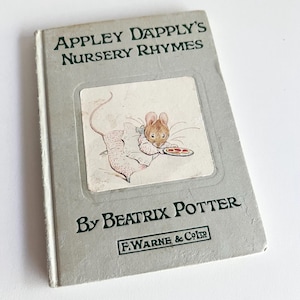 May include: A vintage children's book, "Appley Dapply's Nursery Rhymes" by Beatrix Potter. The cover is pale grey with dark green text. The front features a colour illustration of a mouse holding a plate of food.