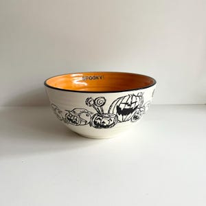 May include: A white ceramic bowl with a black and white Halloween design featuring pumpkins and ghosts. The inside of the bowl is orange.