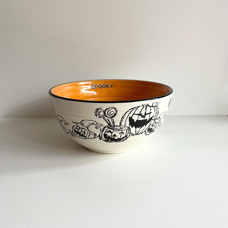 May include: A white ceramic bowl with a black and white Halloween design featuring pumpkins and ghosts. The inside of the bowl is orange.