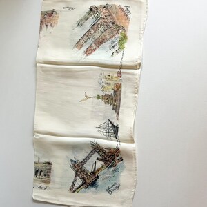 May include: A white cotton tea towel featuring hand-drawn illustrations of iconic London landmarks, such as Tower Bridge, the Houses of Parliament, and the London Eye. The text "London" is written in cursive on the towel.