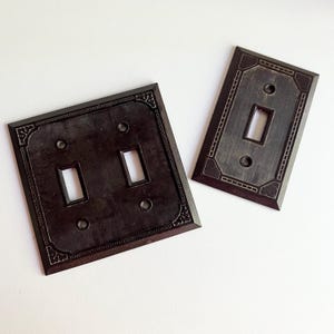 May include: Two dark brown light switch covers with decorative designs. One has two switch openings, the other has one. Both have ornate borders and screw holes. The covers are made of a dark, possibly metallic material, set against a white background.