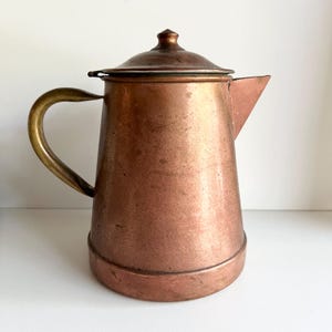Könnte beinhalten: Eine Vintage-Kaffeekanne aus Kupfer mit Messinggriff und Deckel. Die Kanne hat ein klassisches Design mit einem Ausguss zum Ausgießen. Das Kupfer hat einen warmen, rötlich-braunen Farbton, und der Griff ist gebogen. Ein charmantes Küchenutensil.