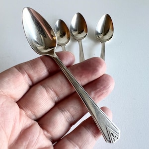 Könnte beinhalten: Ein Satz von vier silbernen Löffeln mit ovalen Schalen und geometrischen Mustern auf den Griffen. Die Löffel sind auf einer weißen Oberfläche angeordnet, wobei sich ein Löffel im Vordergrund und die anderen leicht verschwommen im Hintergrund befinden.