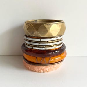 May include: A collection of bangles in a variety of colours and designs. The top bangle is gold-toned with a geometric pattern. Other bangles include white, brown, and orange, with a pink floral-carved bangle at the bottom. A variety of textures and styles.